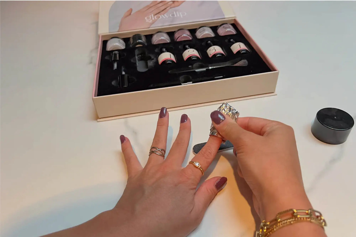 Een paar handen met donkerpaarse nagellak en gouden ringen in een artikel over hoe verwijder je nagellak poeder
