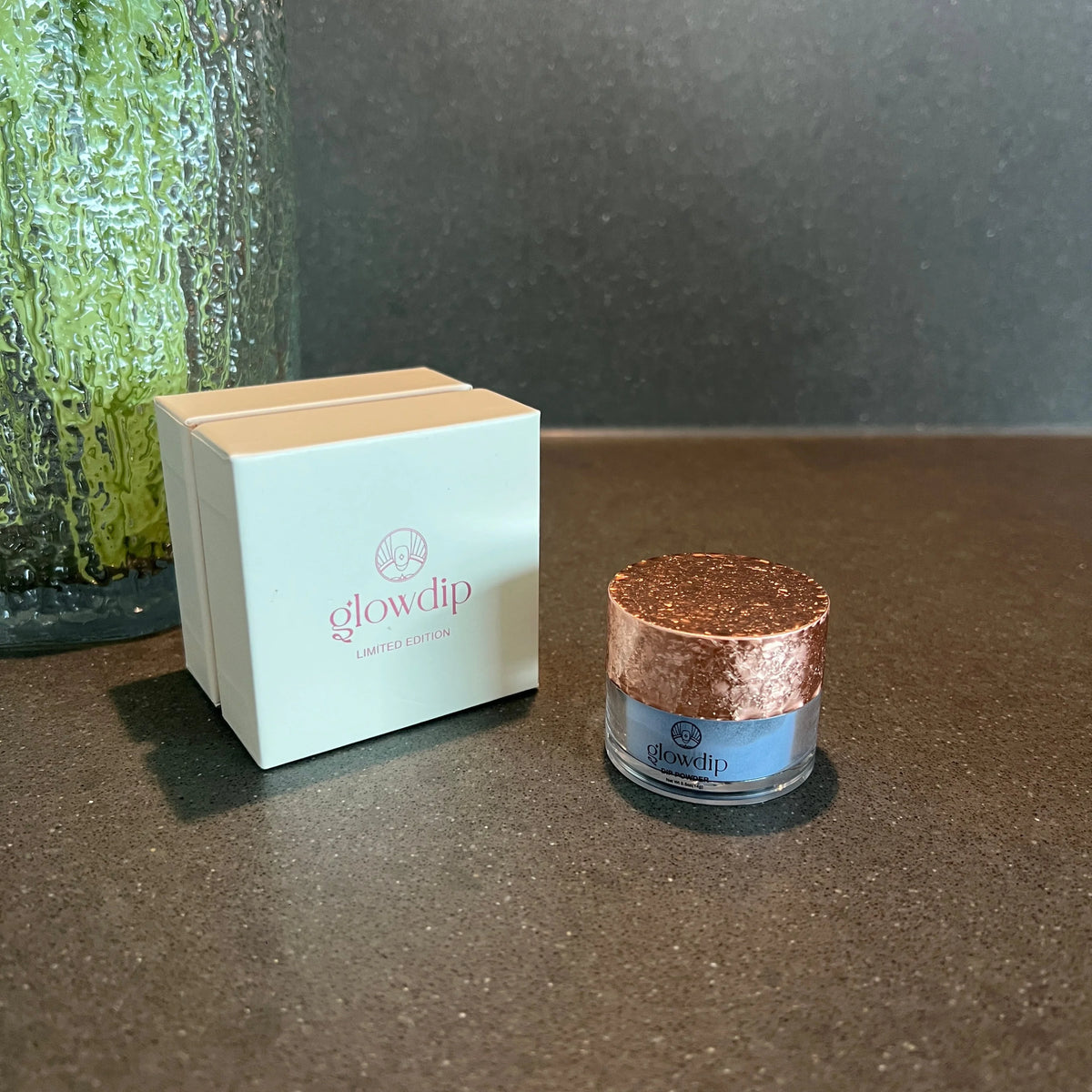 Een glazen pot met lichtblauw moon glow dip poeder en een roségouden glitterdeksel naast een wit merkdoosje